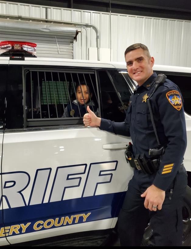 Photo of a young citizen in the back of a Sheriff's Office Tahoe. Sheriff's Office Deputy-Specialist posing for the picture next to the vehicle. 